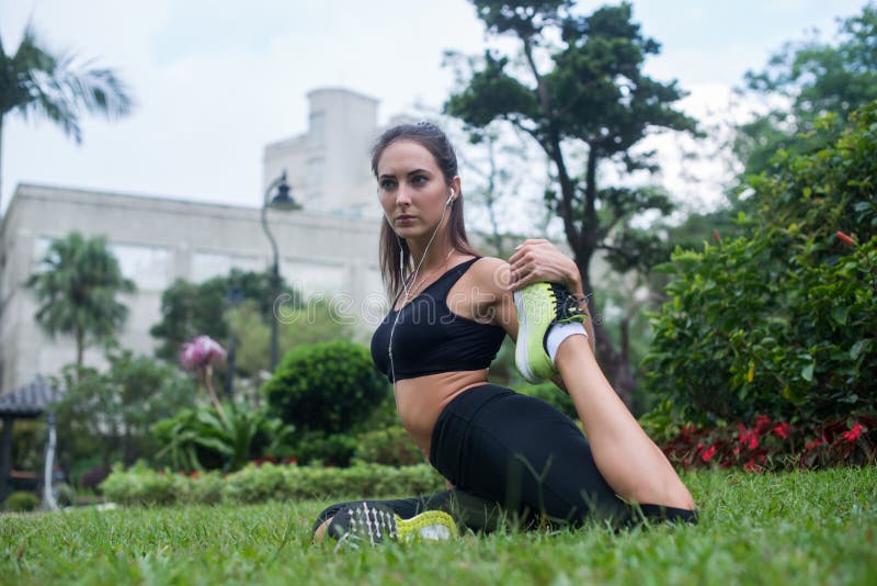 Dysponowana młoda kobieta siedzi na trawie w parku słuchająca ubierał w czarnym sportswear robi rozciągania ćwiczeniu dla nóg pod obraz stock Dysponowana młoda kobieta siedzi na trawie w parku słuchająca ubierał w czarnym sportswear robi rozciągania ćwiczeniu dla nóg pod obraz stock