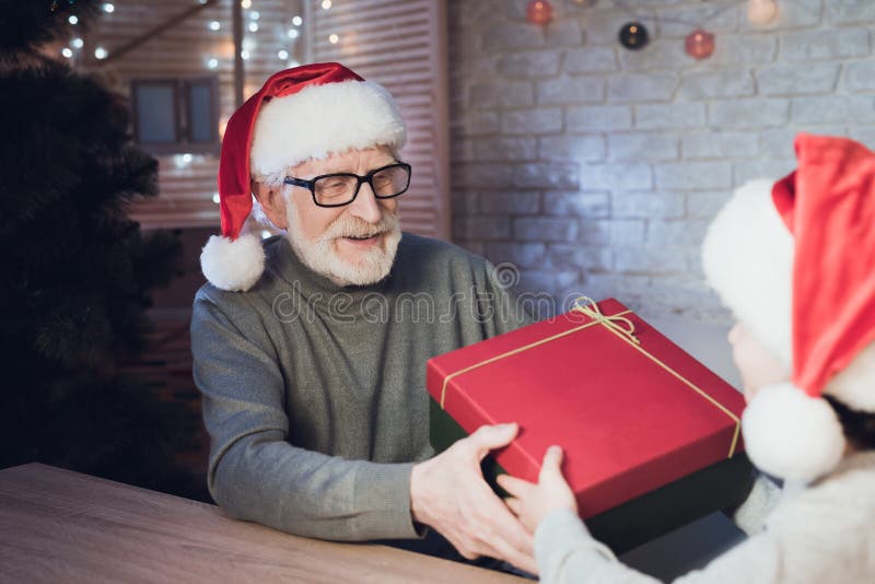 Dziad i wnuk w Święty Mikołaj ` s kapeluszach przy nocą w domu Dziadek daje chłopiec teraźniejszej obraz stock Dziad i wnuk w Święty Mikołaj ` s kapeluszach przy nocą w domu Dziadek daje chłopiec teraźniejszej obraz stock