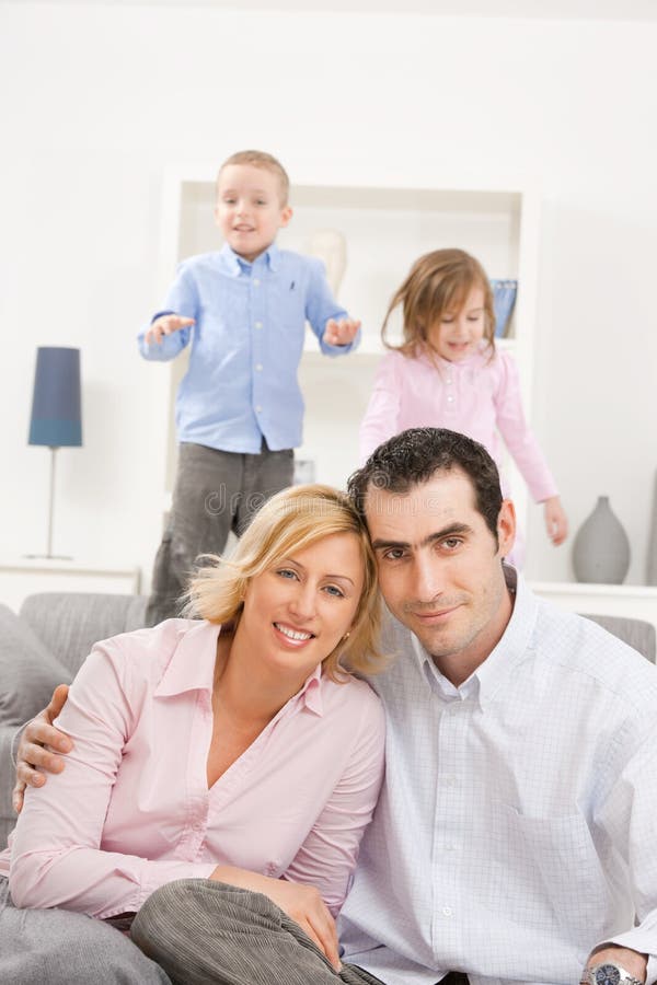 Happy couple sitting at floor at home, embracing. Two children jumping on couch in background. Happy couple sitting at floor at home, embracing. Two children jumping on couch in background.