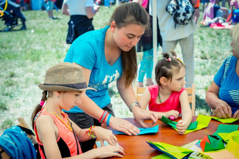 Zaporizhia/Ukraine- June 2, 2018: children and young girl - volunteer at origami workshop, making colorful paper figures on city family charity festival in a city park. Zaporizhia/Ukraine- June 2, 2018: children and young girl - volunteer at origami workshop, making colorful paper figures on city family charity festival in a city park.