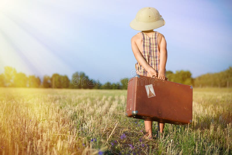 Picture of little boy wearing pith helmet carrying old suitcase in countryside. Backview of kid in plaid romper walking away on sunny flare background. Picture of little boy wearing pith helmet carrying old suitcase in countryside. Backview of kid in plaid romper walking away on sunny flare background.