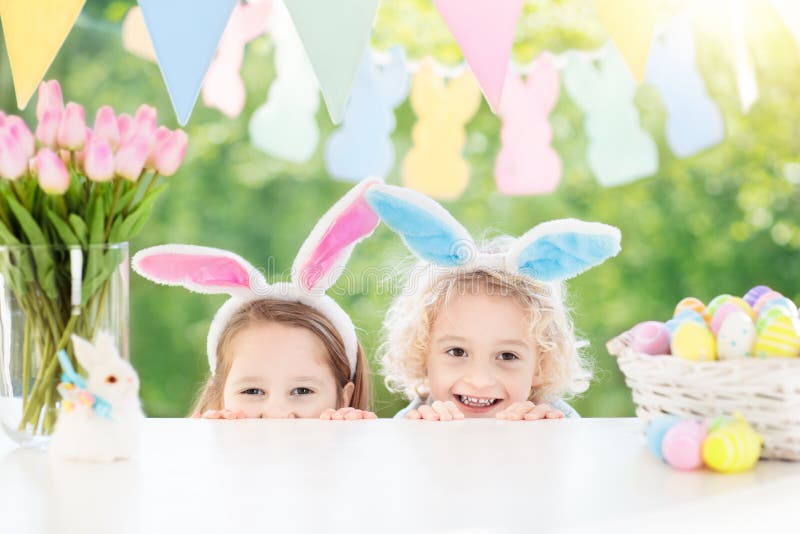 Family Easter morning. Children dye eggs. Kids with bunny ears search for candy and chocolate eggs on Easter egg hunt. Home decoration with tulip flowers, pastel rabbit banner and dye egg basket. Family Easter morning. Children dye eggs. Kids with bunny ears search for candy and chocolate eggs on Easter egg hunt. Home decoration with tulip flowers, pastel rabbit banner and dye egg basket.