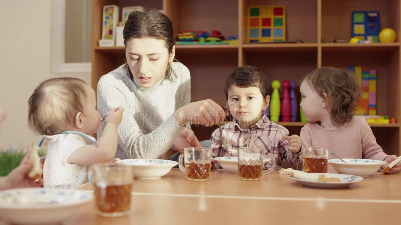 Dziecina nauczyciel siedzi przy stołem z dziećmi i karmi małej ciemnowłosej chłopiec z łyżką inny