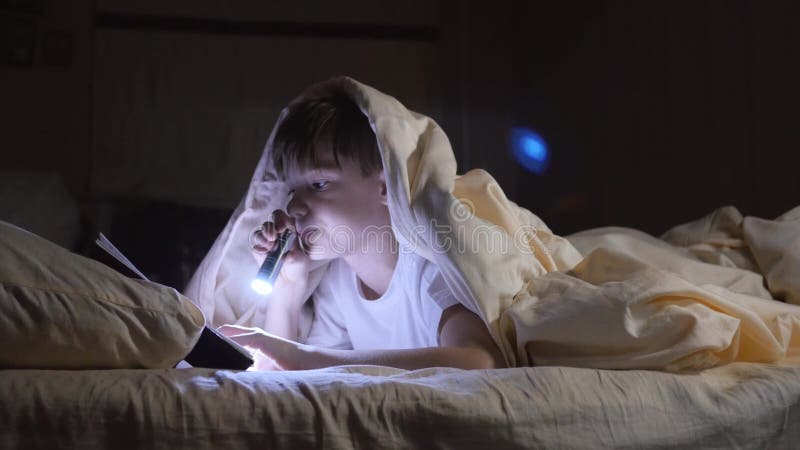 Dziecko czyta książkę pod koc z latarką przy nocą Entuzjastyczna chłopiec