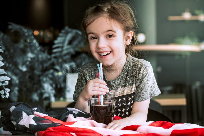 Little girl drinks a drink from a straw in a cafe, the concept of America`s Day and American food. Little girl drinks a drink from a straw in a cafe, the concept of America`s Day and American food