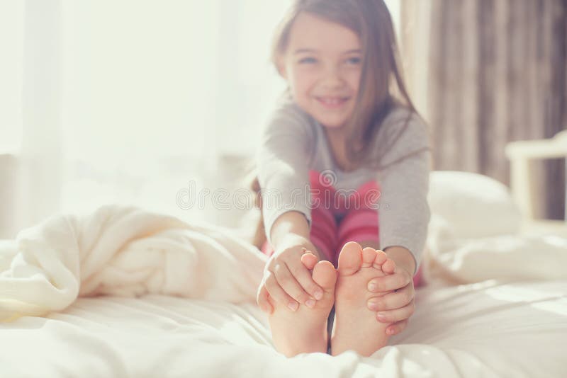 Nice girl,brunette with long uncombed hair,in pink pants and a gray t-shirt,sitting on a large white bed in front of the window,holding hands for feet,waking up early in the morning. Nice girl,brunette with long uncombed hair,in pink pants and a gray t-shirt,sitting on a large white bed in front of the window,holding hands for feet,waking up early in the morning