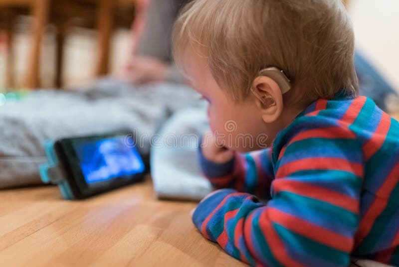 Partially deaf Baby with hearing aid is watching animated tale on smartphone. Partially deaf Baby with hearing aid is watching animated tale on smartphone