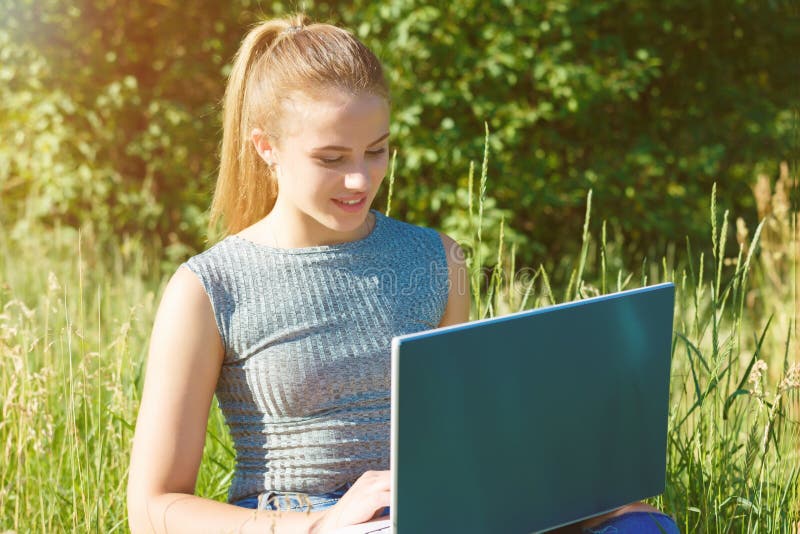 A girl with a laptop in nature among the green grass. Girl with laptop outdoors looking at the screen. A girl with a laptop in nature among the green grass. Girl with laptop outdoors looking at the screen