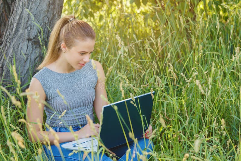 A girl with a laptop in nature among the green grass. Girl with laptop outdoors looking at the screen. A girl with a laptop in nature among the green grass. Girl with laptop outdoors looking at the screen