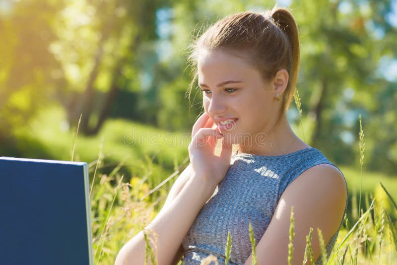 A girl with a laptop in nature among the green grass. Girl with laptop outdoors looking at the screen. A girl with a laptop in nature among the green grass. Girl with laptop outdoors looking at the screen