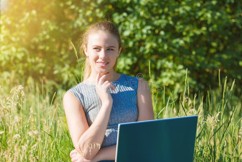 A girl with a laptop in nature among the green grass. Girl with laptop outdoors looking at the screen. A girl with a laptop in nature among the green grass. Girl with laptop outdoors looking at the screen