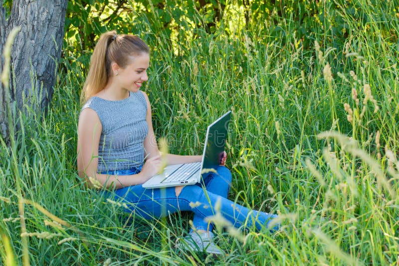 A girl with a laptop in nature among the green grass. Girl with laptop outdoors looking at the screen. A girl with a laptop in nature among the green grass. Girl with laptop outdoors looking at the screen