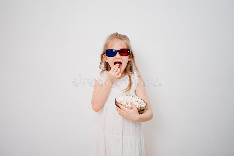 A little girl with glasses to watch 3D movies with a bowl of popcorn stands against a white wall. Home or online cinema. harmful snacking. A little girl with glasses to watch 3D movies with a bowl of popcorn stands against a white wall. Home or online cinema. harmful snacking.