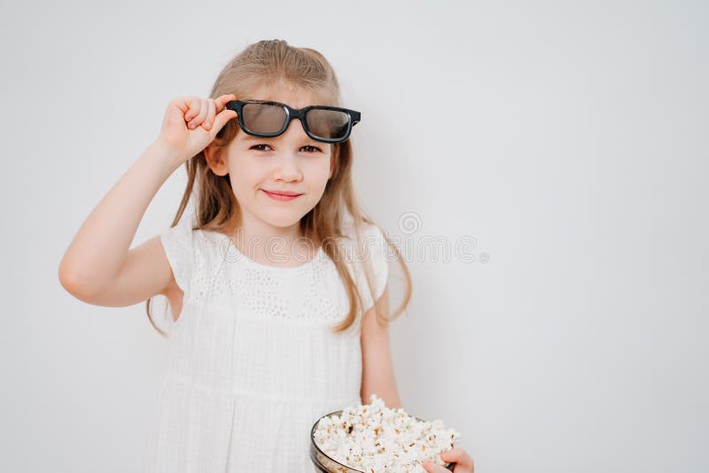 A little girl with glasses to watch 3D movies with a bowl of popcorn stands against a white wall. Home or online cinema. harmful snacking. A little girl with glasses to watch 3D movies with a bowl of popcorn stands against a white wall. Home or online cinema. harmful snacking.