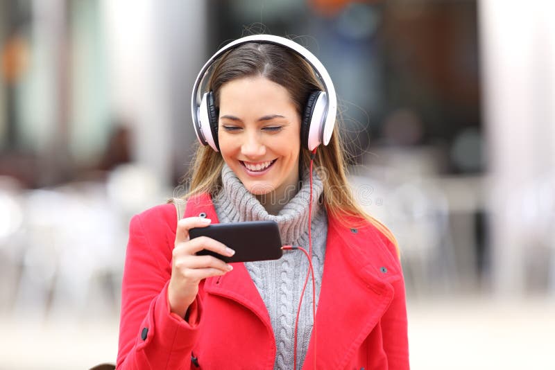 Front view of a happy girl watching videos on line in a smartphone or listening music walking towards camera in the street. Front view of a happy girl watching videos on line in a smartphone or listening music walking towards camera in the street