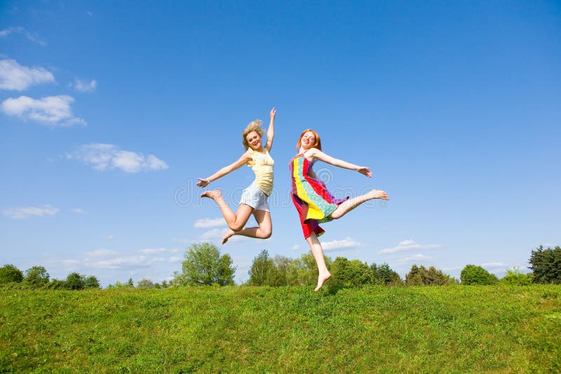 Two happy girls jumping together on green meadow. Against the backdrop of blue sky. Two happy girls jumping together on green meadow. Against the backdrop of blue sky.