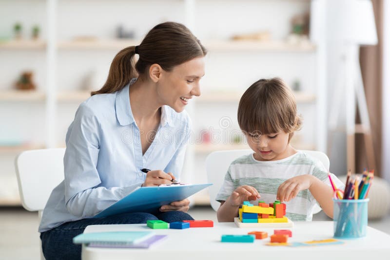 Early kids development. Cute little boy playing logical geometry game, positive woman specialist with notepad watching him at office. Early kids development. Cute little boy playing logical geometry game, positive woman specialist with notepad watching him at office