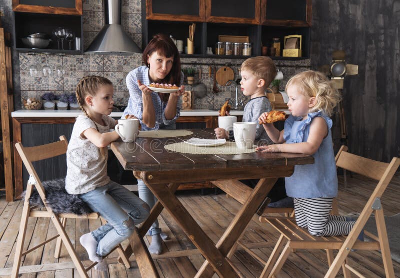 Dîner de famille : Les enfants boivent du thé et la mère apporte un plat de gâteau sur la table photo stock Dîner de famille : Les enfants boivent du thé et la mère apporte un plat de gâteau sur la table photo stock