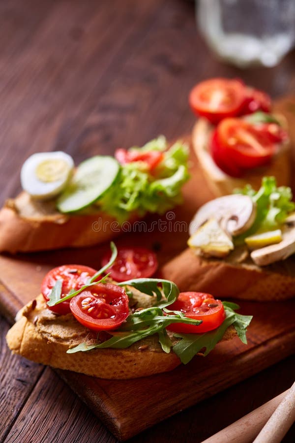 Breakfast sandwich with homemade paste, vegetables and fresh greenson wooden cutting board over rustic background, shallow depth of field, close-up. Delicious morning appetizer. Gourmet bruschetta. Healthy eating concept.