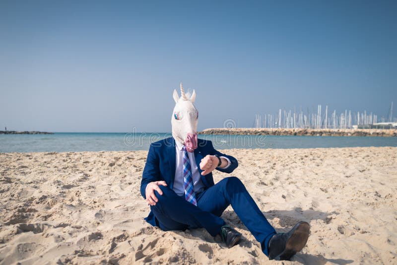 Serious man in comical mask sits on the sand and enjoys sunny day. Unusual guy in suit looking on wristwatch. Strange person relaxing on the beach