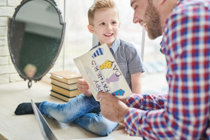 Lovely moments of fatherhood: joyful bearded men reading handmade greeting card given by his little son for Fathers Day while sitting at modern living room. Lovely moments of fatherhood: joyful bearded men reading handmade greeting card given by his little son for Fathers Day while sitting at modern living room