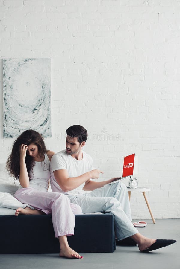 Jovem mulher chateada sentada na cama enquanto o homem aponta com o dedo para o portátil com o site do YouTube fotos de stock