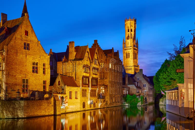 Bruges, Belgium. Evening sunset with blue sky. Water channels of ancient medieval town with view to Belfort van Brugge tower, famous landmark.