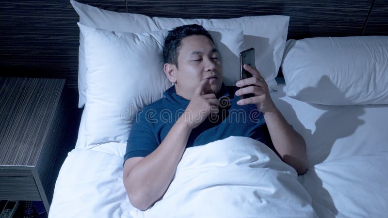 Phone addicted concept, young Asian man using smart phone on bed at midnight, reading chat message, browsing internet, watching movie playing games online porn bedroom lying cell addiction mobile technology dark pillow insomnia alone cellphone sleep male person guy cellular awake smartphone rest face overuse communication evening people tired chronic texting bedtime social sleepy