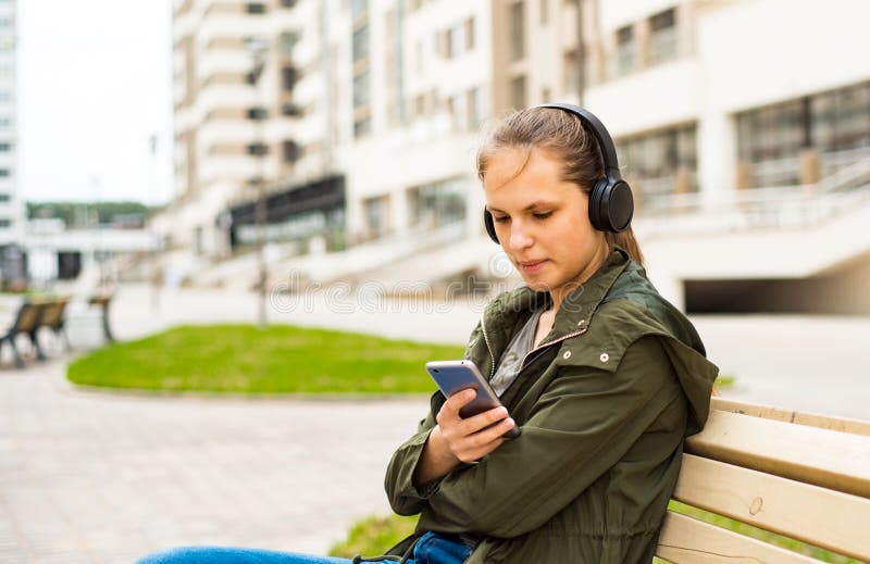 E mujer joven que escucha la m?sica usando el tel?fono m?vil que se sienta en banco fotos de archivo libres de regalías E mujer joven que escucha la m?sica usando el tel?fono m?vil que se sienta en banco fotos de archivo libres de regalías