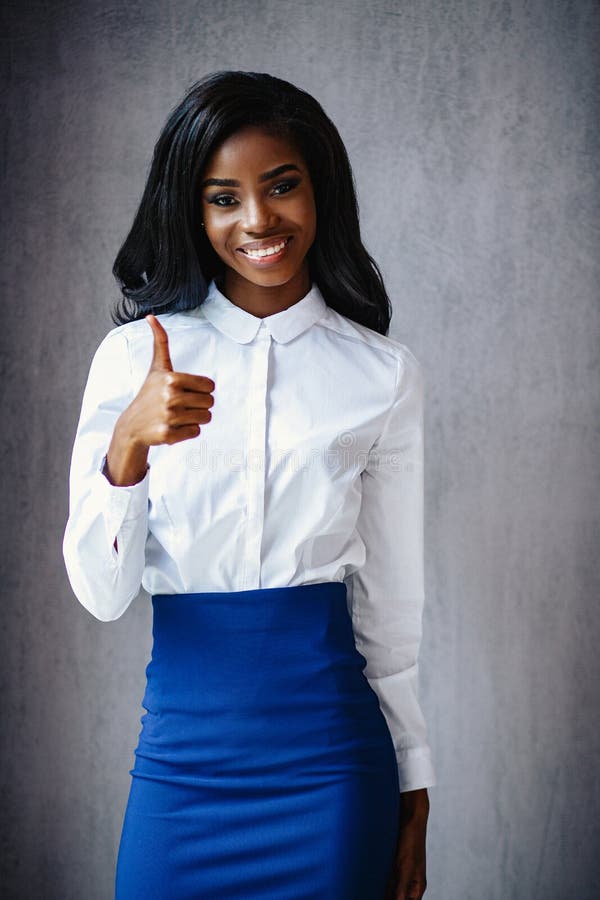 Beautiful African Woman In White Shirt And Blue Skirt Does An Ok Sign With Thumbs Up. Portrait Of African Businesswoman Smiling And Showing Thumb Up. Beautiful African Woman In White Shirt And Blue Skirt Does An Ok Sign With Thumbs Up. Portrait Of African Businesswoman Smiling And Showing Thumb Up.