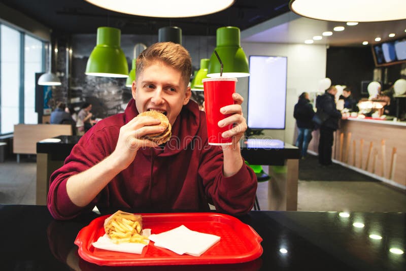 Positive teen in street clothes is eating fast food at a fast-food restaurant, biting a burger, looking at a glass of drink and smiling. Funny student with a fast food tray eating at the restaurant