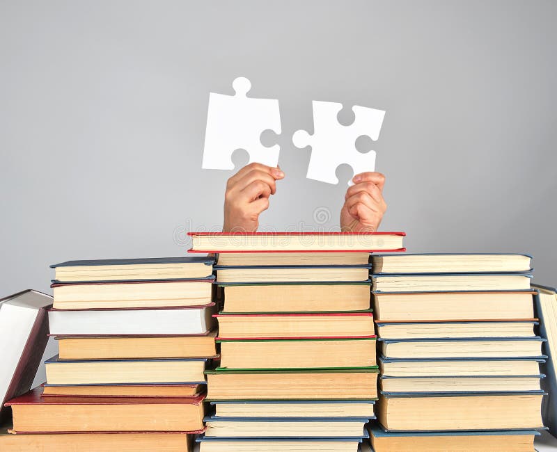 female hands holding big white puzzles over a stack of books, gray background. female hands holding big white puzzles over a stack of books, gray background