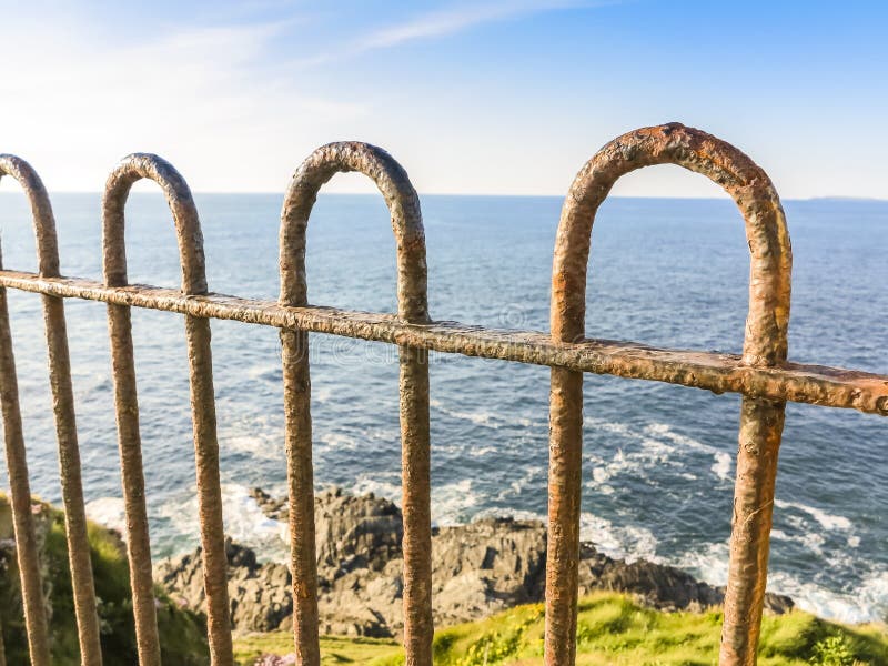 Focus on old rusty fence with rocks and ocean waves below - bright day with old fence in foreground protecting from drop into ocean