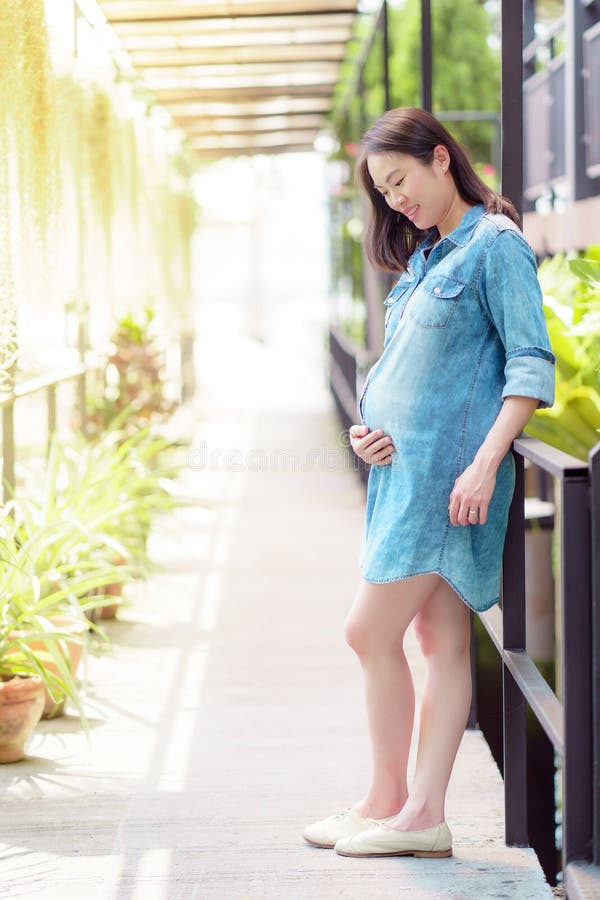 Mulher asiática grávida a usar uma camisa de ganga, de pé no jardim fotos de stock