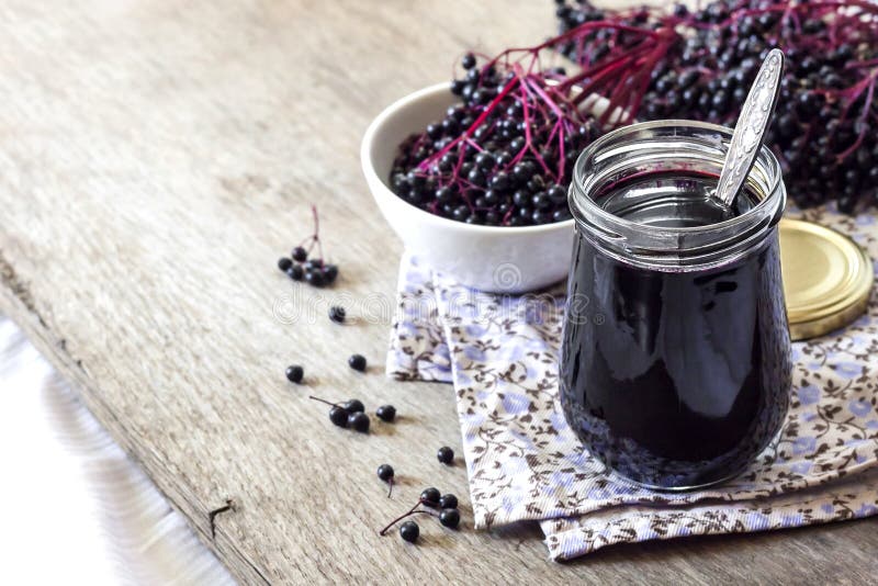 Homemade black elderberry syrup in glass jar and bunches of black elderberry in background, copy space