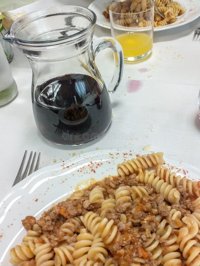 table laden with 2 pasta dishes, a jug of red wine, a glass with orange juice, all placed on a white tablecloth. table laden with 2 pasta dishes, a jug of red wine, a glass with orange juice, all placed on a white tablecloth