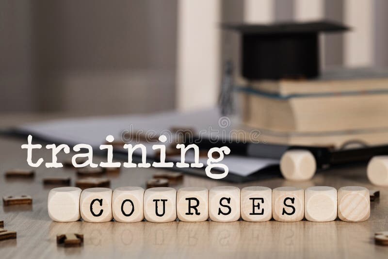 Words TRAINING COURSES composed of wooden dices. Black graduate hat and books in the background. Closeup. Words TRAINING COURSES composed of wooden dices. Black graduate hat and books in the background. Closeup