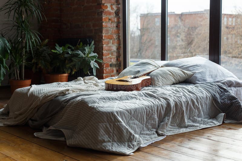 Interior confortável do quarto com cama larga perto das janelas, estilo loft Interior moderno com plantas verdes Guitarra numa ca foto de stock royalty free Interior confortável do quarto com cama larga perto das janelas, estilo loft Interior moderno com plantas verdes Guitarra numa ca foto de stock royalty free
