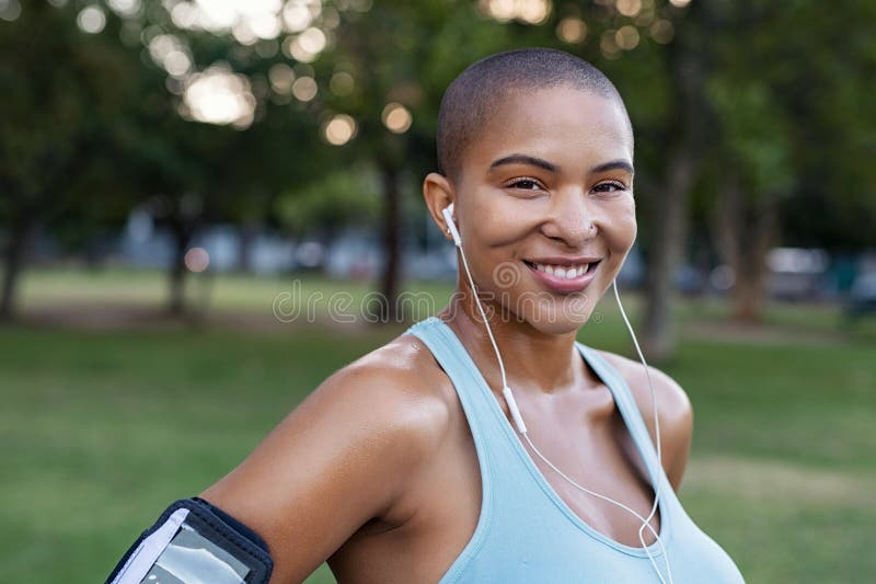 Portrait of a smiling curvy woman listening to music with earphones standing in park after running. Bald young woman looking at camera after fitness workout. Closeup face of satisfied sweating girl after sporty exercises