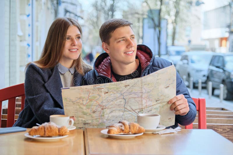 Young tourists man and woman reading map of city in outdoor cafe. Couple drinking coffee tea and eating croissants, spring city background. Young tourists man and woman reading map of city in outdoor cafe. Couple drinking coffee tea and eating croissants, spring city background.