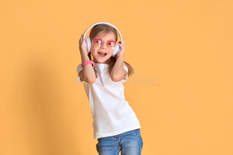 Girl listening music in headphones on yellow background. Cute child in colored sunglasses enjoying happy dance music, smile, posing on pink studio background wall. Girl listening music in headphones on yellow background. Cute child in colored sunglasses enjoying happy dance music, smile, posing on pink studio background wall.