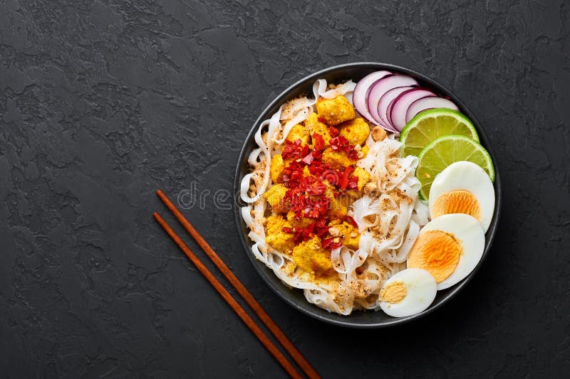 Nan Gyi Thoke in black bowl at dark slate background. Nan gyi Thohk is popular burmese cuisine dish with rice noodles, chicken breasts with spices, eggs, peanuts, lime and red onion. Top view. Nan Gyi Thoke in black bowl at dark slate background. Nan gyi Thohk is popular burmese cuisine dish with rice noodles, chicken breasts with spices, eggs, peanuts, lime and red onion. Top view