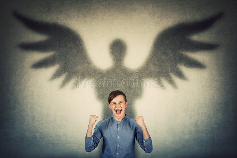Furious teenage boy holding fists up and screaming showing his powers. Confident adolescent guy casting a superhero shadow with angel wings on a dark room wall. Inner strength and ambition concept. Furious teenage boy holding fists up and screaming showing his powers. Confident adolescent guy casting a superhero shadow with angel wings on a dark room wall. Inner strength and ambition concept