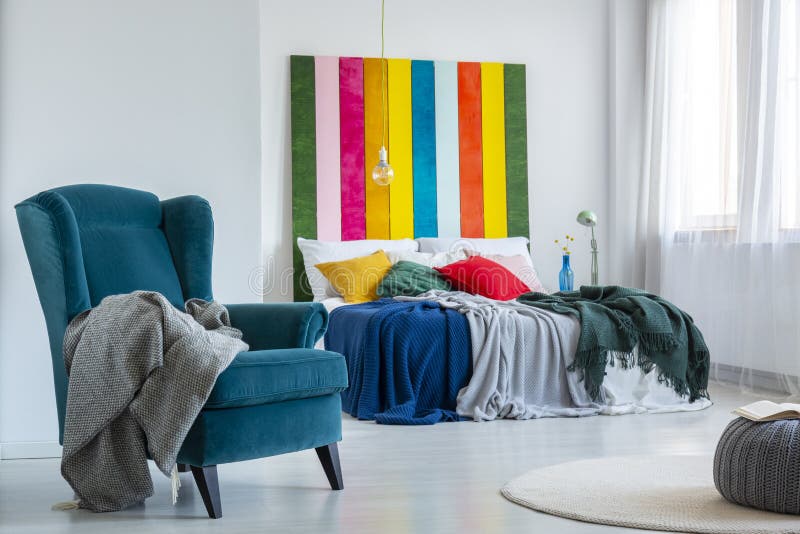 Gray blanket on a comfy, blue armchair next to a colorful bed with cushions in a bright bedroom interior with striped painting on white wall. Real photo. Gray blanket on a comfy, blue armchair next to a colorful bed with cushions in a bright bedroom interior with striped painting on white wall. Real photo