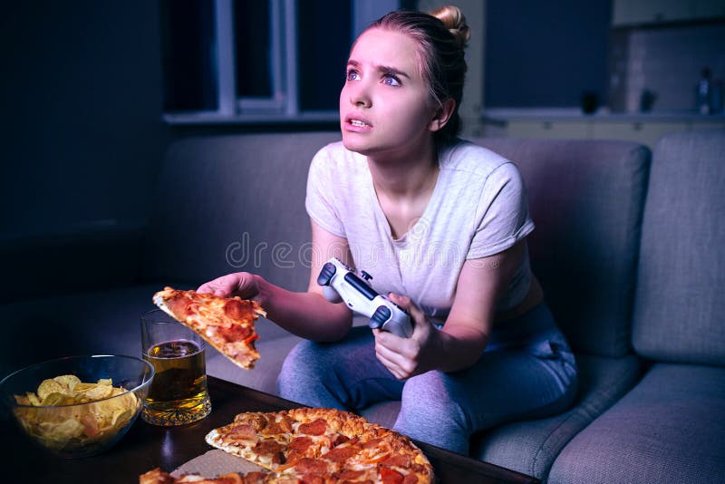Mujer joven que juega al juego en la noche El modelo hambriento mira para arriba en la pantalla Llevar a cabo la parte de la pizz imágenes de archivo libres de regalías Mujer joven que juega al juego en la noche El modelo hambriento mira para arriba en la pantalla Llevar a cabo la parte de la pizz imágenes de archivo libres de regalías