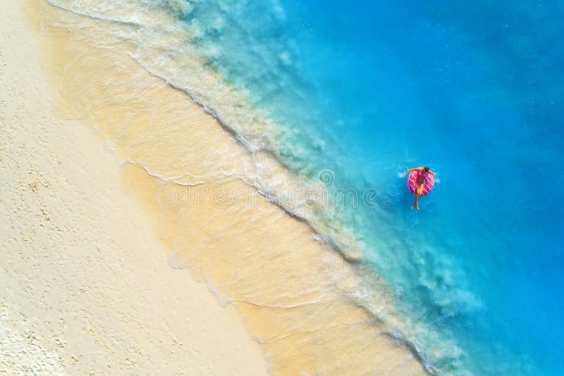 Aerial view of a young woman swimming with the donut swim ring in the clear blue sea with waves at sunset in summer. Tropical aerial landscape with girl, azure water, sandy beach. Top view. Travel. Aerial view of a young woman swimming with the donut swim ring in the clear blue sea with waves at sunset in summer. Tropical aerial landscape with girl, azure water, sandy beach. Top view. Travel