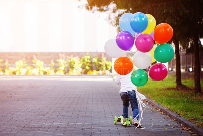 Happy boy playing with a bunch of balloons outside and riding a scooter. Back view. Happy boy playing with a bunch of balloons outside and riding a scooter. Back view.