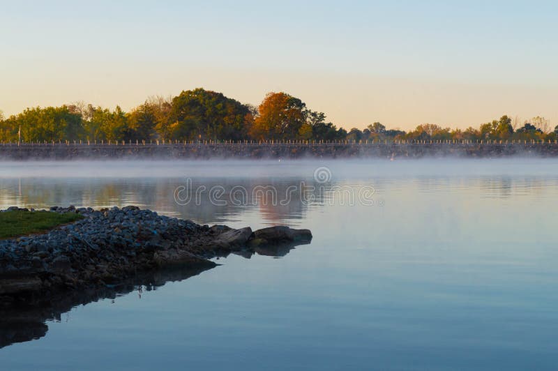 Early morning fog rising from Ohio lake with autumnal colors on trees in background. Early morning fog rising from Ohio lake with autumnal colors on trees in background