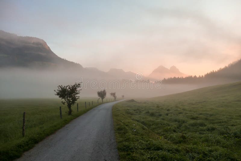 Early morning haze in the Alps. There is a narrow road leading to high mountains through a meadow. The high Alpine peaks