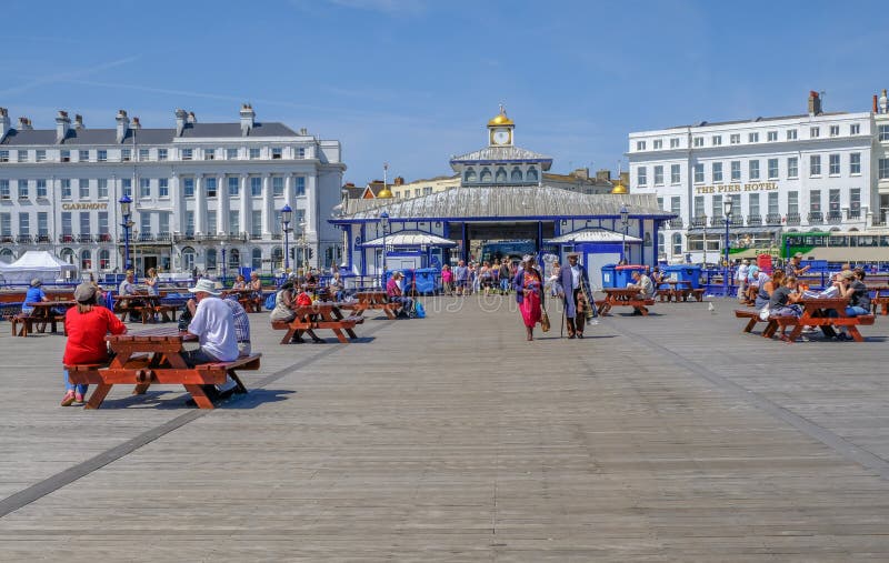Eastbourne Pier, people strolling and sitting on picnic benches on a blue sky summer`s afternoon